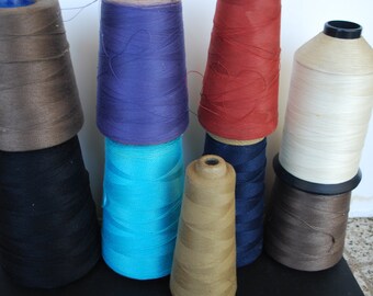 May include: A collection of eight spools of thread in various colors, including brown, black, blue, turquoise, beige, and red. The spools are arranged in a staggered pattern on a dark surface.