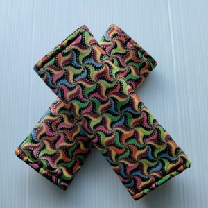 May include: A pair of colorful fabric seatbelt covers with a swirling pattern in shades of green, pink, yellow, and blue on a black background.