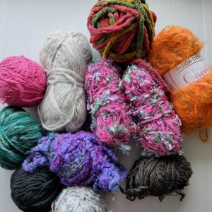 May include: Assortment of yarn in various colors and textures, including a pink, white, green, purple, gray, brown, orange, and multi-colored yarn. The yarn is arranged in a circle on a white surface.