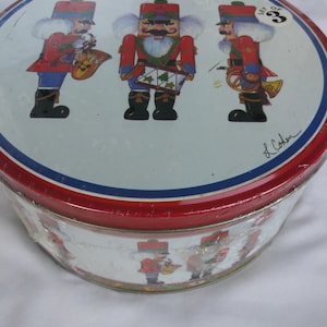 May include: Round tin with a red rim and a lid featuring three nutcracker figures in red and blue uniforms, playing musical instruments. The tin has a white background with a blue and red border. The side of the tin also has nutcracker figures.