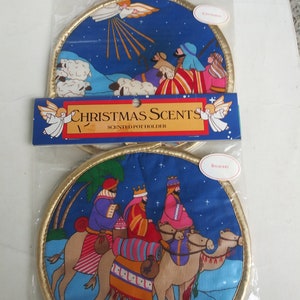 May include: Two Christmas-themed scented pot holders. Each round pot holder features a gold trim and a blue background with a nativity scene. One depicts shepherds and an angel, the other shows the three wise men on camels. The packaging reads "Christmas Scents".