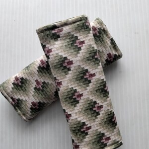 May include: Two rectangular seat belt covers with a geometric pattern in shades of green, beige, and burgundy. The covers are made of a soft, textured fabric and are designed to protect the seat belt from wear and tear.