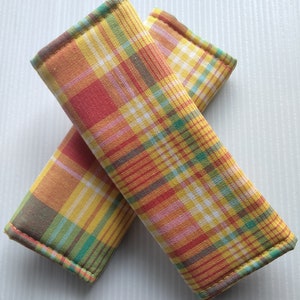 May include: Two colorful seat belt covers with a plaid pattern in shades of yellow, orange, red, and green. The covers are rectangular and padded, designed to provide comfort and style. The edges are neatly stitched.