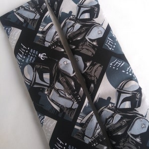 May include: Two rectangular seatbelt covers featuring a repeating pattern of a Mandalorian character in shades of gray, black, and white. The covers have text and a symbol on a black background. The covers are on a white surface.