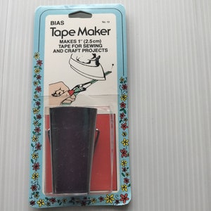 May include: A blue and white package with a red and white floral border. The package contains a silver bias tape maker. The package reads "BIAS Tape Maker" and "MAKES 1" (2.5cm) TAPE FOR SEWING AND CRAFT PROJECTS".