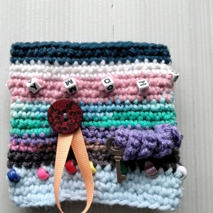 May include: A handmade crochet sensory square in various colors, including blue, pink, and green. It features a decorative button, a small key charm, beads, and lettered beads. A peach-colored ribbon is attached to the button.
