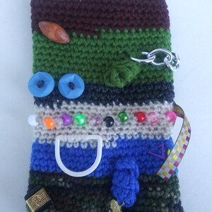 May include: A crocheted pouch with a variety of textures and colors, including brown, green, blue, white, and black. The pouch has a zipper, a key, a button, a wooden bead, and a plastic ring.