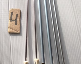 May include: Four silver knitting needles with white plastic tips. The needles are arranged in a row on a white surface. The number "4" is written on a piece of brown paper.