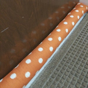 May include: An orange fabric door draft stopper with white polka dots. The cylindrical stopper is placed at the base of a door, resting on a gray patterned rug. The door is a dark brown color.