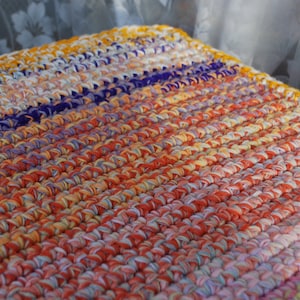 May include: A handmade, rectangular crochet item with a vibrant color palette. The piece features rows of tightly crocheted stitches in shades of orange, yellow, purple, and white. The texture is visible, suggesting a soft, textured surface. The item appears to be a rug or mat.