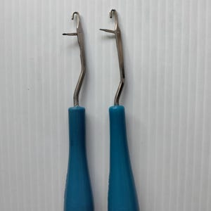 May include: Two crochet hooks with teal blue handles and silver metal hooks. The handles are teardrop-shaped, and the hooks have a curved design. The tools are designed for crafting and textile work.