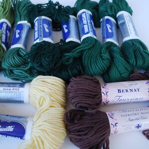 May include: A collection of green, brown, and cream colored yarn skeins. The yarn is labeled "Bernat Tournai Tapestry Wool" and "Bucilla Tapestry Wool".