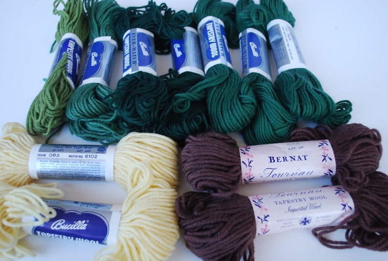 May include: A collection of green, brown, and cream colored yarn skeins. The yarn is labeled "Bernat Tournai Tapestry Wool" and "Bucilla Tapestry Wool".