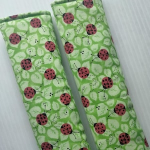 May include: Two green fabric seat belt covers with a repeating pattern of red ladybugs and white leaves. The covers are rectangular and padded, designed to provide comfort and protection.