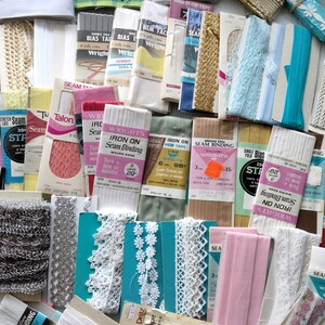 May include: A collection of various sewing notions, including bias tape, seam binding, lace, and ribbon. The items are in a variety of colors, including white, pink, blue, and gold. Packages and spools are visible, with text labels indicating brand names and product details.