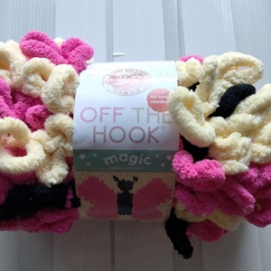 May include: A skein of Lion Brand Off the Hook Magic yarn in pink, cream, and black. The yarn is a chunky, fuzzy texture and is labeled "No tools needed".