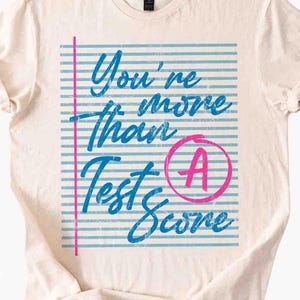 以下が含まれることがあります： クリーム色のTシャツ。青い筆記体で「You're more than Test Score」と書かれています。デザインには、ピンクの縦線とピンクの丸で囲まれた「A」があり、青い線が引かれた背景が特徴です。