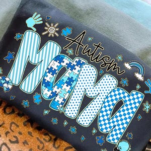 Puede incluir: Camiseta gris oscuro con las palabras "Autism Mama" en un diseño lúdico. La palabra "Mama" está decorada con patrones azules, incluyendo rayas, piezas de rompecabezas y lunares. La palabra "Autism" está en escritura negra.