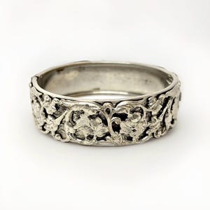 May include: A silver-toned hinged bangle bracelet with intricate floral and leaf carvings. The detailed design wraps around the entire bracelet, creating a textured and ornate appearance. The bracelet is shown against a white background.