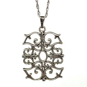 Op de afbeelding: Een zilverkleurige hangerketting met een sierlijk, opengewerkt ontwerp. De hanger heeft een rechthoekige vorm met krullende bloemen- en geometrische patronen en een ovale midden. De ketting is een delicate, gedraaide stijl.