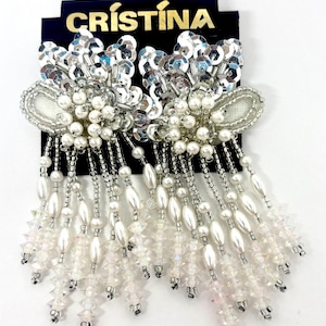 May include: A pair of elaborate, dangling earrings. The earrings feature a cluster of silver sequins and white faux pearls. Long strands of white beads and crystals hang from the bottom. The earrings are displayed on a black card with the name "CRISTINA" in gold lettering.