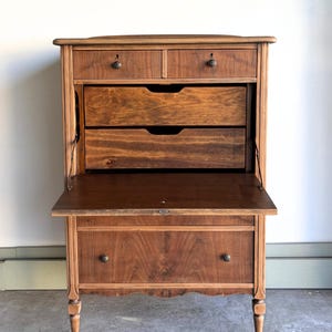 May include: A brown wooden secretary desk featuring two small top drawers with round metal knobs, two open interior shelves, a fold-down writing surface, and one large bottom drawer with two round metal knobs, supported by four turned legs.