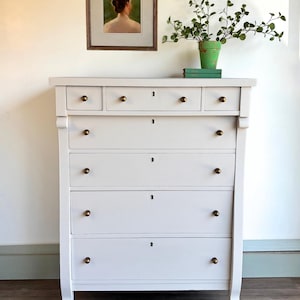 May include: A tall, light gray dresser with six drawers and brass knobs. The top section has smaller drawers. A framed painting hangs above the dresser, and a green potted plant sits on top. The dresser has a classic, elegant design.