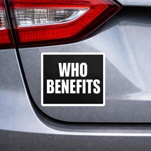 May include: A rectangular black and white sticker with the words "WHO BENEFITS" in bold, white capital letters. The sticker is attached to the rear of a silver car, near the rear light.