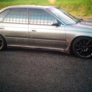 Puede incluir: Un sedán plateado con llantas negras estacionado sobre una superficie pavimentada. El coche tiene cristales tintados con un estampado blanco y negro. La parte delantera del coche es de un tono plateado más oscuro. El coche está estacionado sobre una superficie pavimentada.