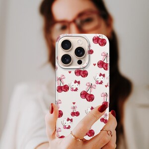 May include: A white phone case featuring a pattern of red cherries, pink bows, and cartoon cat characters. The case has a clear border and covers the phone's camera lenses. The design includes small red hearts scattered across the surface.
