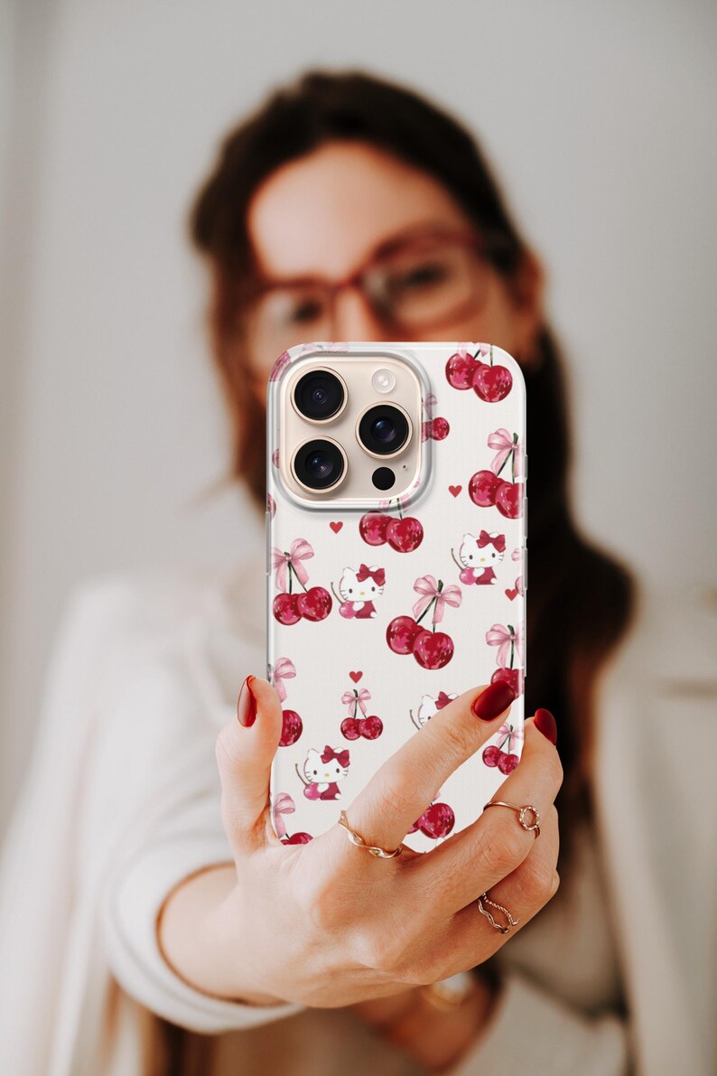 May include: A white phone case featuring a pattern of red cherries, pink bows, and cartoon cat characters. The case has a clear border and covers the phone's camera lenses. The design includes small red hearts scattered across the surface.