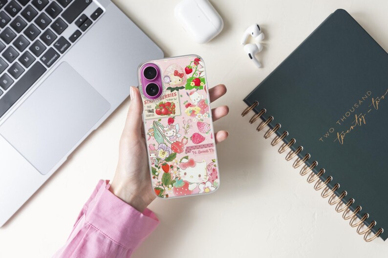 May include: A clear phone case with a pink background featuring a strawberry and floral design. The case has a collage of images, including strawberries, flowers, and a cartoon character. The text "Strawberries" is visible on the case.