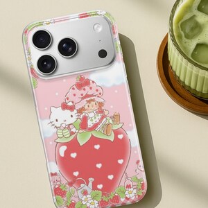 May include: A pink phone case featuring a whimsical design with Hello Kitty and a girl character sitting on a large strawberry. The case has a clear border and a light pink background with white clouds and strawberry details.