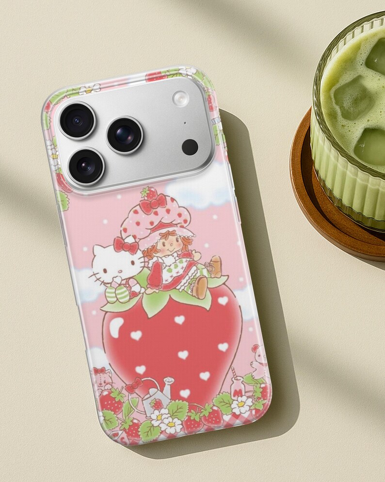 May include: A pink phone case featuring a whimsical design with Hello Kitty and a girl character sitting on a large strawberry. The case has a clear border and a light pink background with white clouds and strawberry details.