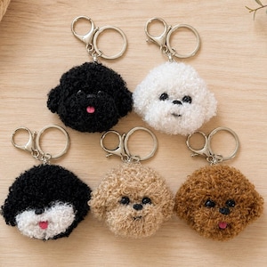 May include: Five plush dog head keychains with silver-toned metal rings and lobster clasps. The collection includes a solid black, a solid white, a black and white, a tan, and a brown dog, all featuring fuzzy textures, black eyes, and pink tongues.