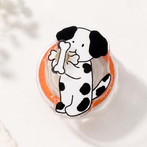 May include: A white and black cartoon dog with black spots holds a bone. The dog is on a clear, round base with an orange border. The background is white with a sprig of white flowers.