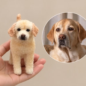 May include: A small, needle-felted cream-coloured dog figurine with a textured wool finish, dark eyes, and a brown nose, displayed alongside a circular inset photograph of a real yellow Labrador retriever with similar facial features.