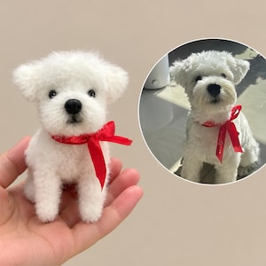 May include: A small, white, needle-felted dog figurine with a black nose and eyes, wearing a bright red satin ribbon tied in a bow around its neck. Beside it is a circular inset photo of a real white dog wearing an identical red ribbon.