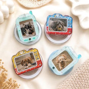 May include: Four circular phone grips featuring decorative frames shaped like vintage cameras and mobile phones. Each frame displays a photograph of a different dog breed. The frames are colored in light blue, red, and yellow with printed icons.