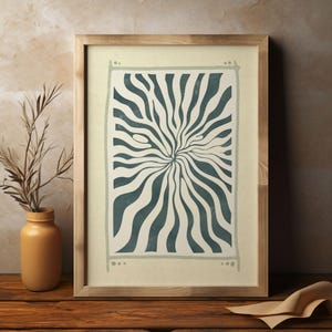May include: Abstract art print in a wooden frame. The artwork features a teal and cream-colored design with a central burst pattern. The frame is a light brown color. A yellow vase with dried plants is to the left of the frame.