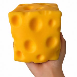 Könnte beinhalten: Ein leuchtend gelber, quaderförmiger Käseblock mit mehreren großen, unregelmäßigen Löchern. Der Käse wird in einer Hand vor weißem Hintergrund gehalten, wodurch seine Textur und Farbe hervorgehoben werden.