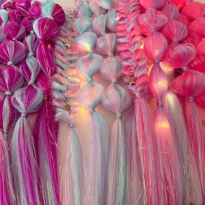 Può includere: Primo piano di extension per capelli intrecciati colorati nelle tonalità del rosa, viola e blu. Le trecce sono adornate da piccole luci, creando un effetto luminoso. I capelli sono acconciati con un design a treccia a bolle.