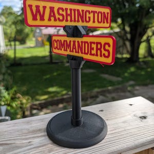 May include: A tabletop sign featuring two rectangular yellow plaques with red borders and red lettering. The top plaque reads WASHINGTON and the bottom plaque reads COMMANDERS. The signs are mounted on a black vertical pole and a round black base.