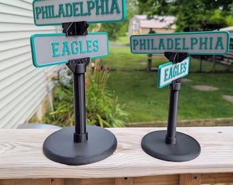 Pode incluir: Duas réplicas de placas de rua impressas em 3D com o texto Philadelphia e Eagles em letras verde-azuladas sobre fundo branco com bordas pretas. Cada placa é montada num poste cilíndrico preto com uma base circular.