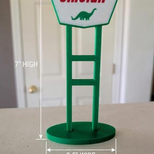 May include: A green and white Sinclair gas station sign replica featuring a green dinosaur graphic and the text Sinclair. The sign stands on an oval base and measures 7 inches high by 3.5 inches wide as indicated by the visible measurement markers.