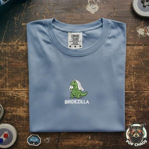 Puede incluir: Camiseta azul claro con cuello redondo. Presenta un dinosaurio verde con un velo blanco, con la palabra "BRIDEZILLA" debajo. La camiseta es de la marca "Comfort Colors".
