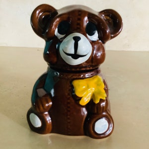 May include: A brown ceramic teddy bear figurine with a yellow bow tie. The bear has black eyes and a white nose and mouth.