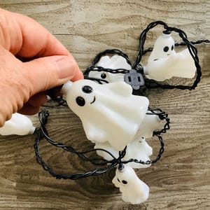 May include: A string of decorative holiday lights featuring multiple white, translucent ghost-shaped covers with black eyes and smiling mouths. The ghosts are attached to a thin black electrical cord, which includes a small plastic plug connector.