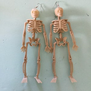 May include: Two beige plastic skeleton earrings with silver-colored ear wires. Each earring features a full skeleton design, including a skull, rib cage, arms, legs, and feet. The earrings are displayed on a light blue surface.