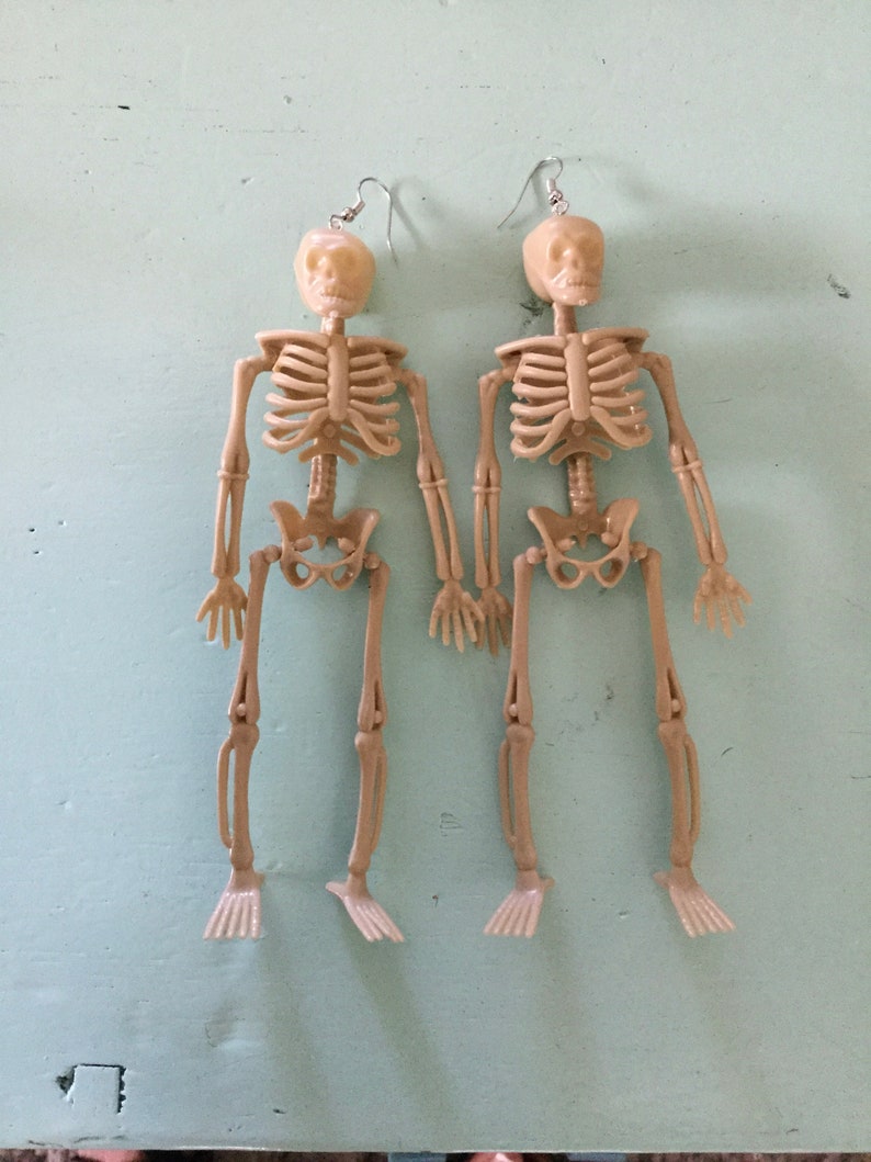 May include: Two beige plastic skeleton earrings with silver-colored ear wires. Each earring features a full skeleton design, including a skull, rib cage, arms, legs, and feet. The earrings are displayed on a light blue surface.